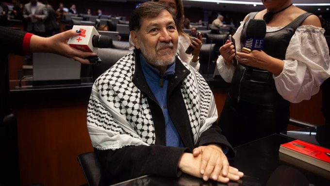 Gerardo Fernandez Norona, an older man with a beard and glasses, sits at a desk wearing a black and white checkered kufiya over a blue jacket, hands clasped in front. A woman in a black off-shoulder top holds a microphone toward him from the left, with a red manicured hand holding a white box nearby. Another woman with long hair stands to the right holding a microphone. The setting is an indoor conference room with wooden walls, chairs, people in the background, and a book on the desk.