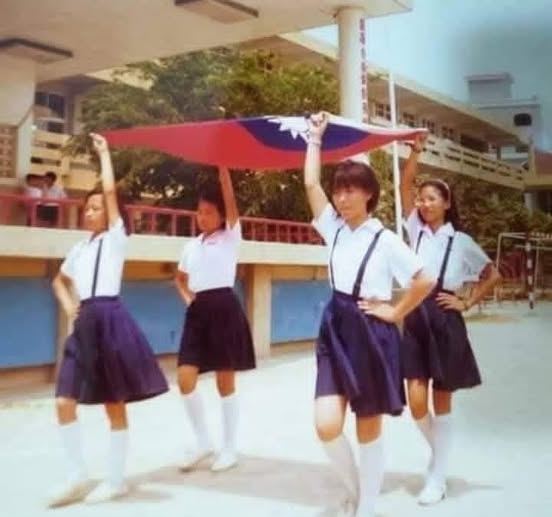 Four young girls in white short-sleeve shirts with black suspender straps over navy blue pleated skirts and white socks stand on a schoolyard pavement holding up a large red flag with a blue square and white sun emblem at the top of a tall white flagpole under a covered walkway with green trees and school buildings in the background during what appears to be a flag ceremony