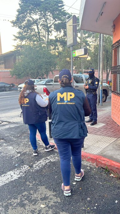 First image shows multiple agents in dark blue jackets with yellow MP logos and police vests standing on a nighttime urban street lined with buildings, some with blue and green walls, under streetlights, some faces blurred, holding items like rifles. Second image depicts a group of agents in MP jackets and vests gathered near a building entrance at night, with blurred faces, one holding a phone, urban setting with poles and lights. Third image features agents in dark uniforms near a blue building and orange traffic barrel on a dimly lit street at night, faces blurred, standing in a line. Fourth image displays two agents, one male in uniform and one female in blue vest with MP logo, standing near a parked SUV and a wall with a sign, in daylight urban area with trees and poles.