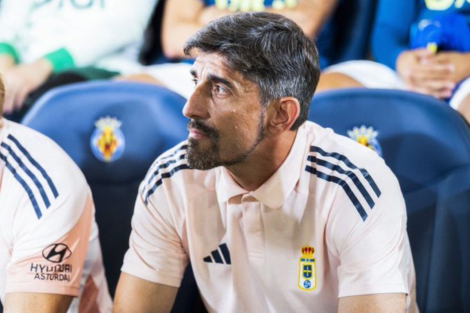 Predrag Paunovic sits in stadium seats wearing a pink Getafe polo shirt with Adidas stripes and team crest on chest, gray hair and beard, looking sideways pensively, surrounded by blue seats and other people in similar attire including a bottle nearby.