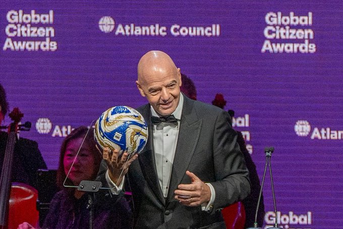 Gianni Infantino, a bald man in a black tuxedo with a white shirt and bow tie, smiles while holding a large blue and gold decorative soccer ball in his right hand on a stage. Purple backdrop displays Global Citizen Awards and Atlantic Council logos. Microphones and a violinist with a cello are visible nearby. A smartphone on a tripod stands beside him.
