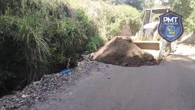 A roadside construction site features a yellow bulldozer loading dirt from a large pile onto its bucket, positioned next to a narrow ditch with rocks and a blue plastic sheet partially covering it. Lush green vegetation and tall grasses line the edge of the dirt road. Blue and white PMT shields are overlaid on the image corners.