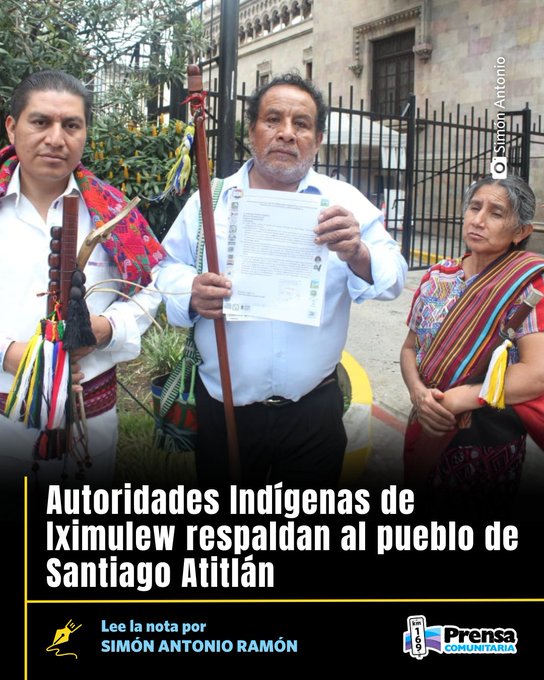Three indigenous individuals in traditional embroidered clothing stand outdoors near a metal fence and plants, with one man holding a document titled Autoridades IndÃ­genas de Iximulew respaldan al pueblo de Santiago AtitlÃ¡n, another man holding a staff with multicolored ribbons, and a woman standing beside them; text overlay includes Lee la nota por SIMON ANTONIO RAMÃN and Prensa Comunitaria logo.