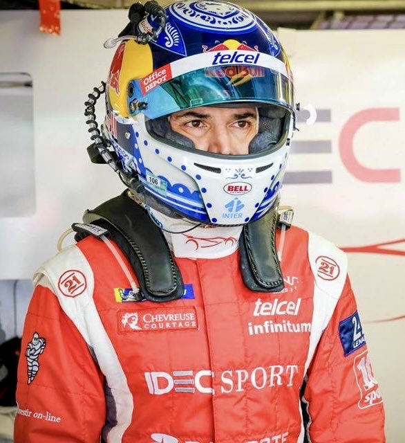 A male driver with short dark hair visible through the open visor of a blue helmet featuring Red Bull and Telcel logos stands in front of a red wall. He wears a red and white racing suit with sponsor patches including Chevrolet, Telcel, and Infinitum, and a black neck brace. The helmet has yellow accents and a clear visor with a rearview mirror cable.