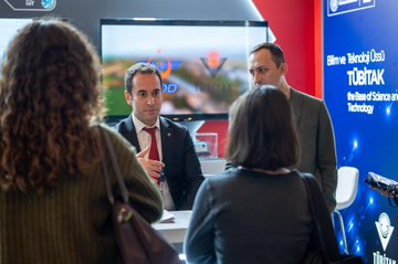 First image shows a group of professionals including a woman in green attire gesturing while speaking to two men in suits at a modern booth with TÜBİTAK logos, display screens showing technology interfaces, glass cases with exhibits, and a QR code banner in a spacious indoor congress hall with hanging lights and upper levels visible. Second image depicts a man in a suit with red tie standing and gesturing near a table at a booth with a large screen displaying aerial drone footage, surrounded by women and men in business attire, TÜBİTAK branding on blue and red panels, and English text The State of Science and Technology. Third image captures several young men in casual and formal wear gathered around a laptop screen at a booth with TÜBİTAK logo projections, digital panels showing tech interfaces, and red and gray booth design in a lit exhibition space with windows. Fourth image features multiple people in suits and casual clothes standing and interacting near booth displays with screens, chairs, and TÜBİTAK signage on white and red structures in a large hall with high ceilings and adjacent exhibit areas.