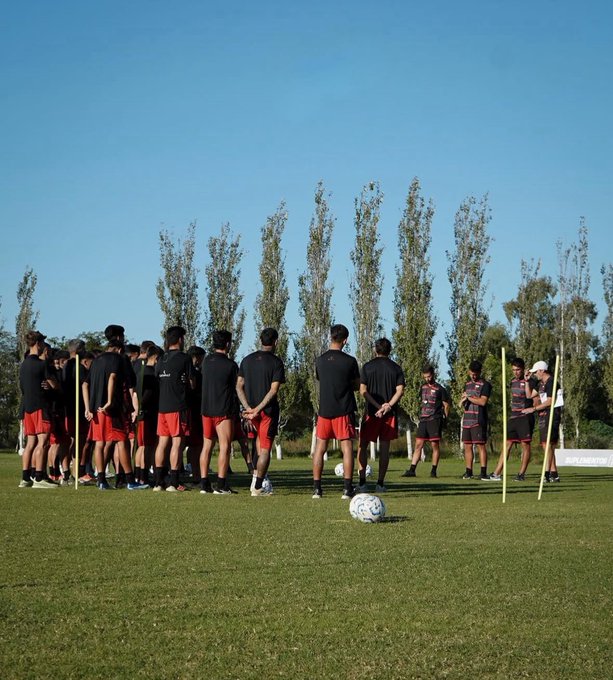 Group of male football players in black jerseys and red shorts stand in a line on a green grass field during training, facing forward with hands behind backs, under a clear blue sky with tall green trees in the background, yellow training poles set up nearby, and a white soccer ball on the ground in front of them.