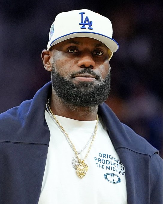 LeBron James wears a white Los Angeles Lakers cap with blue lettering, a gold chain necklace featuring a heart-shaped pendant, and a navy blue jacket over a shirt. His facial hair includes a beard and mustache. The background shows a blurred indoor arena setting with blue and purple lighting.