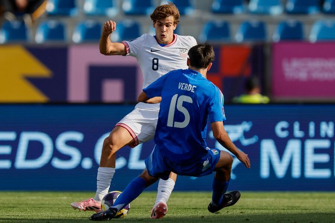 Two soccer players in action on a green field during a match, one wearing a white jersey with number 9 for Estados Unidos pushing the ball forward while another in a blue jersey with number 15 for Italia attempts to tackle from behind, surrounded by stadium seating and advertising banners including Clines and Meds.