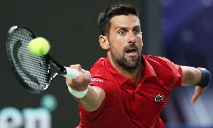 Novak Djokovic in red Lacoste polo shirt and wristband mid-serve hitting yellow tennis ball with black racket over net on indoor court with dark background and green padding.