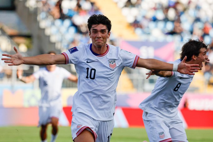 Two male soccer players in white USA national team jerseys with numbers 10 and 8 celebrate a goal by raising arms wide on green field during match against Italia stadium with crowd blurred background other players visible in distance.