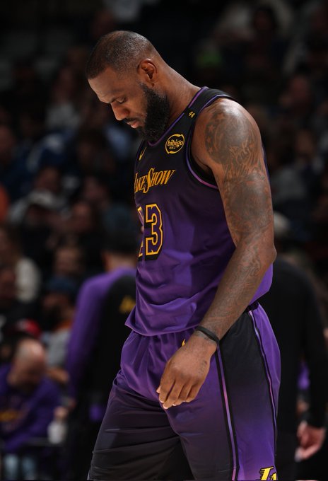 LeBron James stands on a basketball court wearing a purple Los Angeles Lakers jersey with number 23 and gold accents, black shorts, wristband, and tattoos visible on his arms, with a blurred crowd in the background and arena lighting.