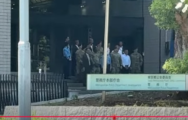 Several men wearing dark suits and light blue uniforms stand in a group on concrete pavement outside a modern gray building with large windows and a sloped entrance. Green leafy trees and a metal fence are visible to the right. A white sign mounted on the building reads 警視庁 in large black characters with additional Japanese text below indicating Tokyo Metropolitan Police Department and substation details. A streetlight pole stands nearby under a clear blue sky.