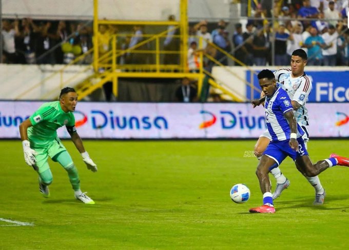 Imagen de un partido de fútbol en un estadio con gradas llenas de aficionados ondeando banderas y sosteniendo carteles, jugadores en uniforme azul y blanco de Honduras presionando hacia la portería defendida por un guardameta en verde de Costa Rica que se lanza para bloquear un tiro, balón en movimiento cerca de la línea de gol con marcas publicitarias como Diunsa visibles en las vallas laterales y un marcador electrónico en el fondo.