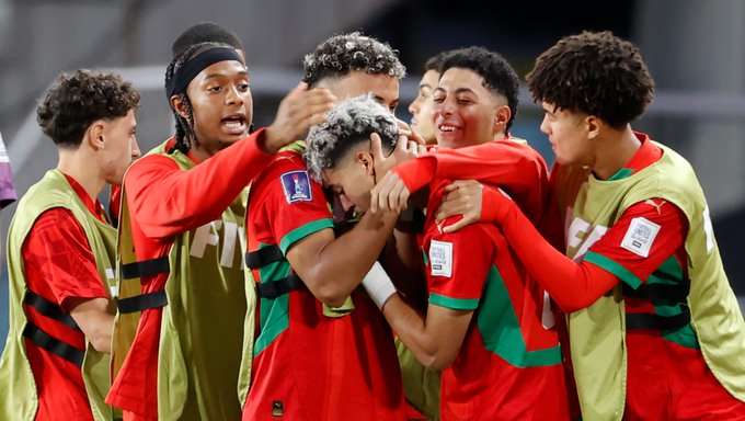 Group of young male soccer players in red jerseys with green accents and the Moroccan flag emblem, wearing headbands and arm sleeves, huddled together in celebration on a green field, some with arms around shoulders and hands on heads, surrounded by yellow training bibs on the ground, stadium seating in background.