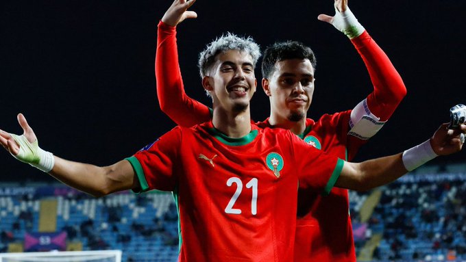 Two young male soccer players in red Morocco national team jerseys with green accents and number 21 on one celebrate a goal by raising arms joyfully on a stadium field at night with blue seats in background.