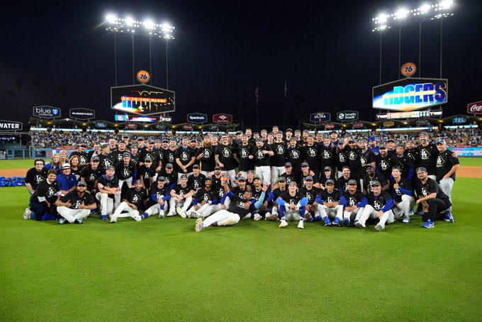 Group of Los Angeles Dodgers baseball players in white and blue uniforms posing together on green field at Dodger Stadium at night, illuminated by bright stadium lights, with large video screens displaying sponsor logos like Michelob Ultra in background, crowd visible in stands.