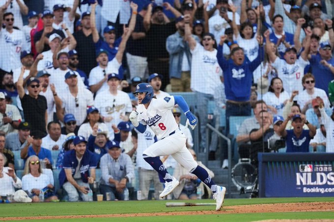 Un jugador de beisbol con uniforme blanco y azul de los Dodgers, numero 8 en la espalda, corre hacia la primera base en un estadio lleno de fanaticos vestidos de blanco y azul que levantan los brazos celebrando, con el marcador NLDS visible en una pantalla y el campo de juego verde de fondo.