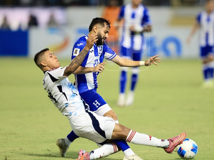 Two soccer players in blue and white uniforms compete for the ball on a green field during a match, one player tackling from behind while the other advances with the ball near their foot, surrounded by teammates in similar jerseys and a stadium crowd in the background under stadium lights.