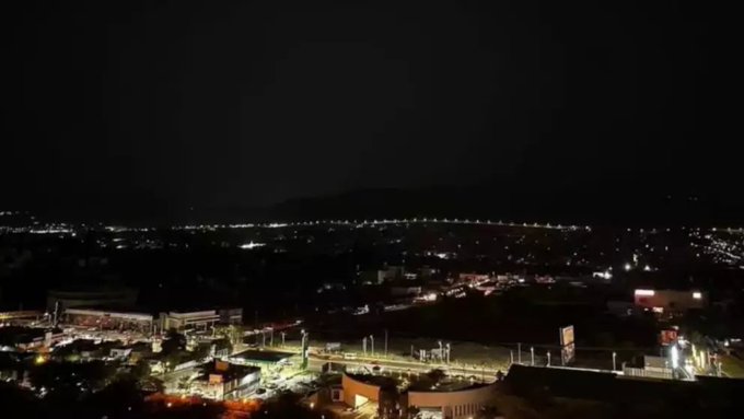 Vista aérea nocturna de una zona urbana en expansión con numerosos edificios y calles iluminadas por luces dispersas y faros de vehículos, una colina prominente en el fondo bajo un cielo oscuro, que indica áreas de corte de energía en medio de las luces de la ciudad.