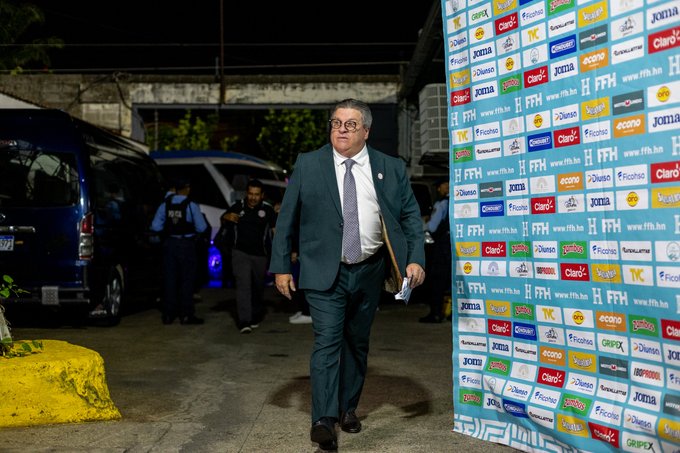 Man in dark suit white shirt and tie holding folder walks near sponsor banner wall with logos like Coca-Cola and Adidas at nighttime outdoor event security personnel in background vans and yellow barrier present.
