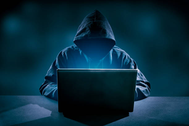 Hooded figure working on a laptop in a dark room, representing cybercrime