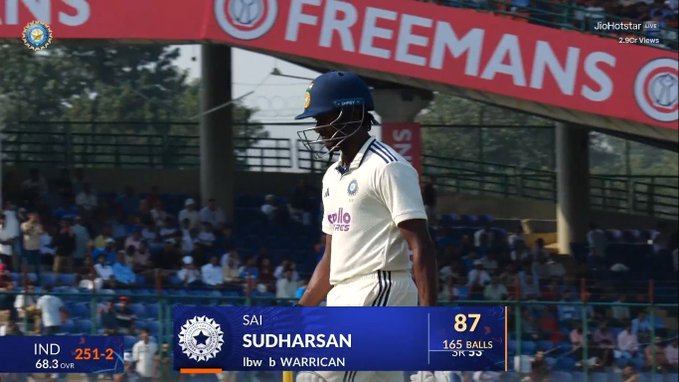 Sai Sudharsan in white cricket uniform and blue helmet walks off the field towards the pavilion with a dejected expression, scoreboard in foreground shows SAI SUDHARSAN 87 off 165 balls, background includes stadium seating with spectators and a red banner reading FREEMANS above sponsor logos.