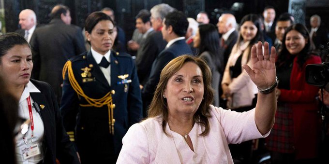Dina Boluarte, una mujer de cabello castaño que viste una blusa rosa claro, se encuentra en un evento formal lleno de gente levantando su mano derecha en un gesto, rodeada de diversos asistentes con trajes, uniformes militares con trenzas doradas y vestidos, algunos con cámaras, en un ambiente interior con paneles de madera.