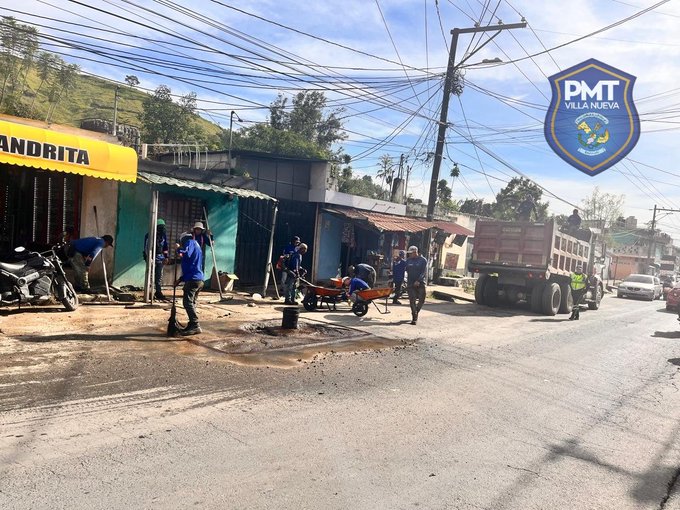 Several uniformed officers in blue shirts and pants stand and work on a dirt road with potholes, using shovels and a wheelbarrow filled with gravel next to a yellow dump truck loaded with dirt. Small green and yellow buildings with signs including one reading ANDRITA line the street, with motorcycles parked nearby and power lines overhead. A clear blue sky with some clouds is visible, and a PMT shield logo appears in the corner.
