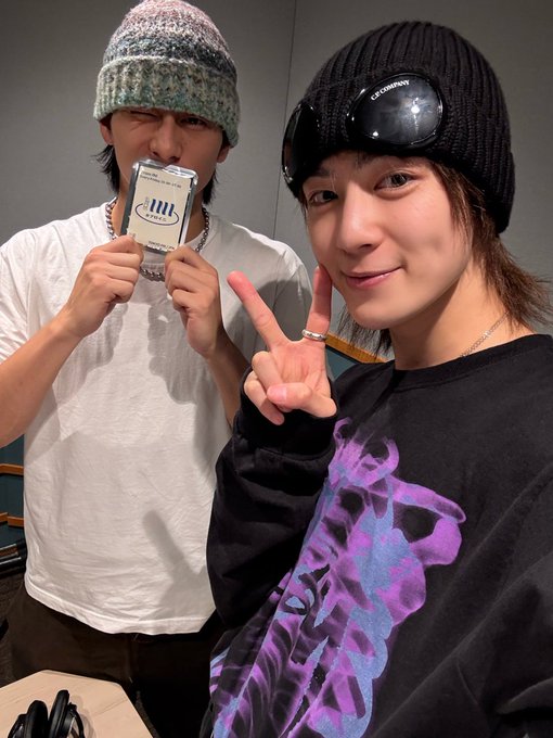 Two young men posing indoors near a table with shoes on it. The one on the left wears a white t-shirt, holds a small white rectangular object like a snack package near his mouth, has a chain necklace with INI pendant, and makes a peace sign with his right hand. He has short dark hair and wears light pants. The one on the right wears a black hooded sweatshirt with purple flame-like graphics, black beanie, black sunglasses, has shoulder-length dark hair, smiles, and makes a peace sign with his left hand. He stands with hands in pockets of dark pants.