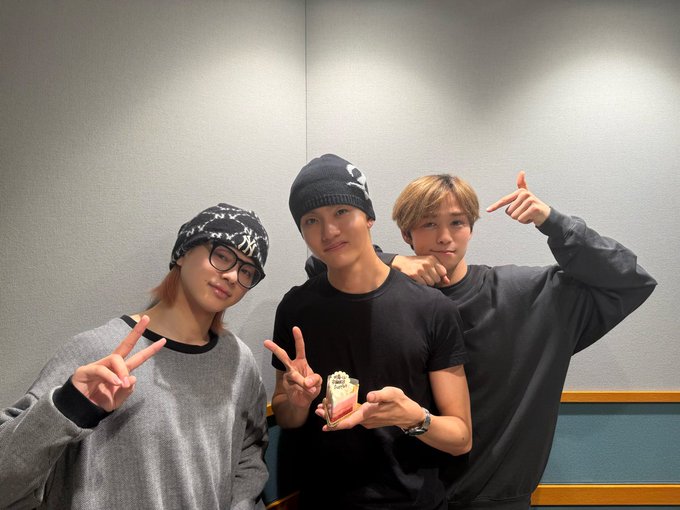 Three young men in casual streetwear pose together in a studio setting with neutral walls and lighting. The left man wears a gray hoodie with black trim, glasses, and a black beanie, making a peace sign with his right hand. The center man in a black t-shirt holds a small white cake with pink frosting and a candle, smiling. The right man in an oversized gray hoodie points forward with his right hand, wearing a black beanie. They stand close, appearing celebratory.