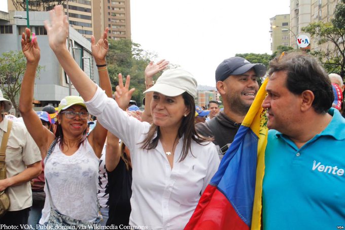 Grupo de personas en un mitin político al aire libre en un entorno urbano con edificios altos y árboles de fondo. Mujer central con blusa blanca y gorra levanta su mano derecha, sonriente, identificada como María Corina Machado del contexto post. Los partidarios de los alrededores incluyen hombres y mujeres con atuendos informales, algunos con gorras y bolsas amarillas, levantando la mano con entusiasmo. Prominente bandera colombiana amarilla, azul y roja sostenida por un hombre con camisa azul. Otra mujer con top colorido y gorra a la izquierda. Ambiente de celebración con multitud diversa.