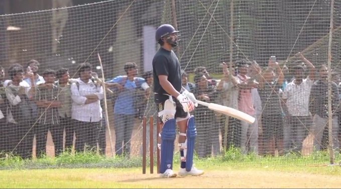 Indian cricketer Rohit Sharma stands at the batting crease in practice nets on a grassy outdoor field. He wears a black helmet with visor, black short-sleeve shirt, white batting gloves, blue leg pads, and holds a wooden cricket bat in his right hand while resting it on the ground. His stance is relaxed yet prepared, with blue trousers tucked into white shoes. Behind the net fence, a diverse crowd of men in casual shirts and pants watches attentively, some standing and others leaning forward. The scene conveys high interest in his practice session.