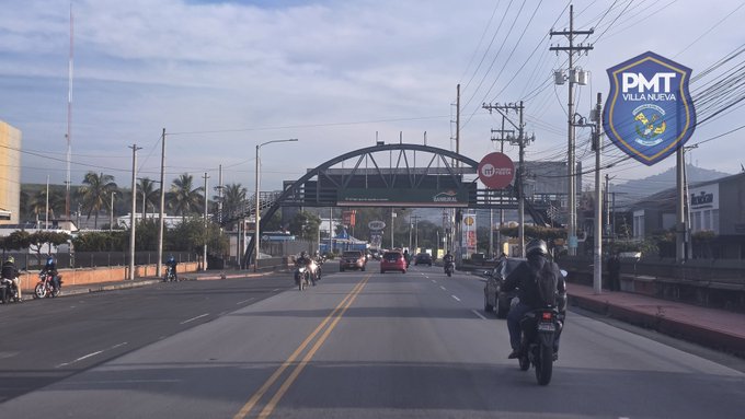 Urban street scene at Bulevar El Frutal in zona 5 with a large arched structure overhead displaying signage, multiple lanes of asphalt road with yellow center lines, several motorcycles ridden by officers in blue uniforms and helmets, cars and other vehicles in traffic, palm trees along the sides, utility poles with wires, and a stop sign visible near the arch.