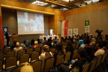 İlk görüntüde dört erkek panelist masa başında oturuyor, Türk bayrakları ve su şişeleri önünde, arka planda ekran ve duvar panelleri görülüyor. İkinci görüntüde konferans salonunda çeşitli yaş ve cinsiyetten katılımcılar sıralı sandalyelerde oturuyor, bazıları not alıyor, Türk bayrakları ve su bardakları masalarda, arka planda Yeşilyurt tabelaları asılı. Üçüncü görüntüde bir konuşmacı kürsüde sunum yapıyor, ekran üzerinde video oynatılıyor, izleyiciler başörtülü kadınlar ve erkekler dahil oturuyor, Türk bayrakları ve yeşil logolar duvarlarda. Dördüncü görüntüde grup fotoğrafı, çeşitli cinsiyetten ve etnik kökenlerden 20den fazla kişi ayakta poz veriyor, arka planda ekran ve Yeşilyurt tabelası, bazıları başörtülü ve takım elbiseli.