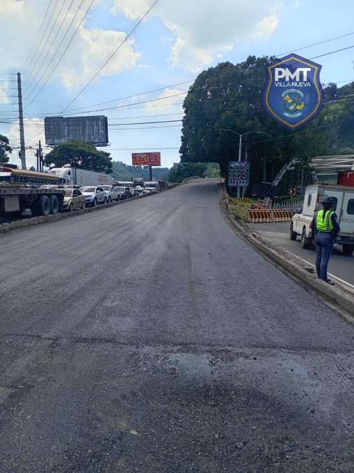 A paved road curves uphill under a partly cloudy sky with power lines overhead and billboards in the background. Several vehicles including trucks and cars are parked or stopped along the roadside near orange traffic cones and barriers. A white police vehicle with emergency lights is positioned near the cones, and a person in a high-visibility vest stands nearby. A large blue and yellow shield emblem labeled PMT is mounted on a pole beside a tree, overlooking the scene.