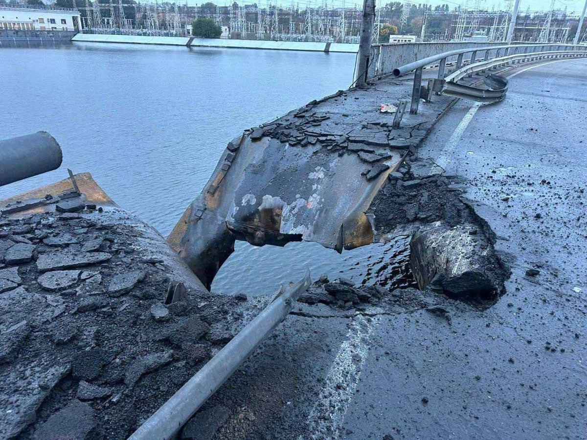 一座横跨水体的桥梁因明显的撞击而严重损坏，大部分道路倒塌并摇摇欲坠地悬挂在边缘，烧焦和发黑的金属碎片散落在周围，栏杆弯曲和折断，周围地区包括建筑物和船只在阴云密布的天空下在背景中可见。