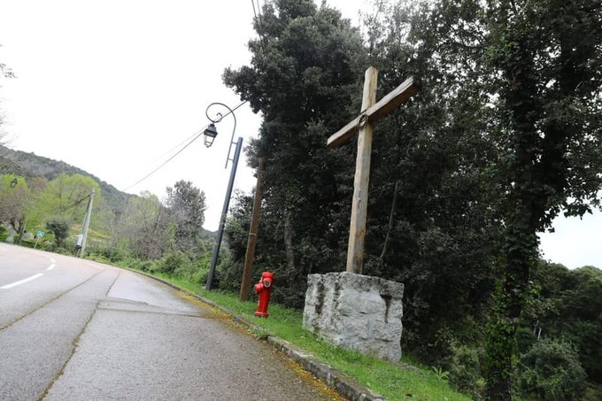 A tall wooden cross mounted on a stone pedestal stands beside a rural road surrounded by green trees and bushes under a cloudy sky with a red fire hydrant nearby and utility poles along the roadside in a scenic hillside setting