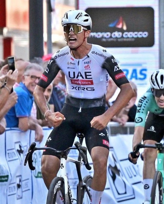 A male cyclist in white helmet with visor sunglasses and UAE Team Emirates black and white jersey pumps fists in celebration while riding a white road bike with black handlebars during a race on a sunny urban street lined with barriers and spectators some holding phones older man in blue shirt nearby another cyclist in green kit behind sponsor logos on barriers like Movistar and Kern visible.