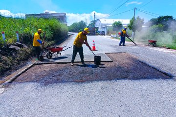 First image shows three workers in yellow vests and helmets on a roadside with green vegetation and buildings, one pushing a wheelbarrow, another spreading asphalt from a bucket with a shovel, and a third using a leaf blower, with traffic cones and a fresh asphalt patch on the road. Second image depicts a group of workers in yellow vests around a red truck unloading materials, spreading asphalt on a road patch using a rake, surrounded by greenery and a clear sky. Third image features a yellow roller compacting a fresh asphalt patch on the road, with workers in vests nearby, green bushes, and a COVIAL logo on the vehicle. Fourth image displays a completed asphalt repair on a rural road with traffic cones, workers in the background, lush green hills, and scattered buildings under a blue sky.