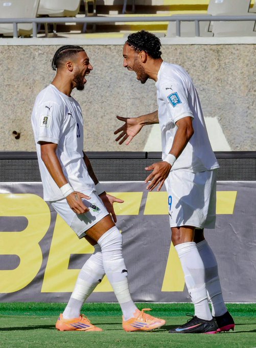 Dos jugadores de fútbol con uniformes blancos de Puma con el número 9 en una camiseta se paran en un campo verde cerca de los asientos amarillos del estadio, riendo y gesticulando animadamente el uno hacia el otro, vistiendo tacos naranjas y negros, con un fondo borroso de gradas y una pancarta de patrocinador.