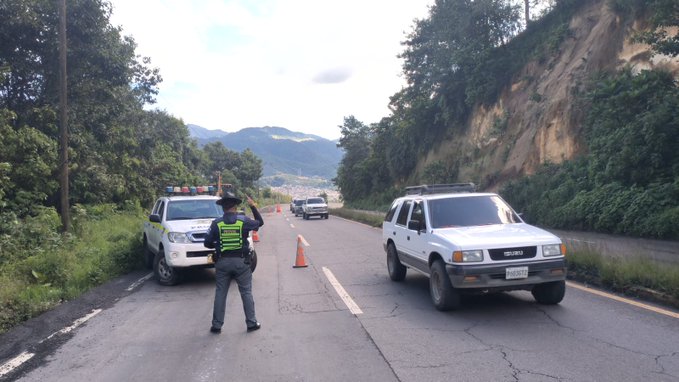 Un oficial de Provial con chaleco reflectante amarillo y sombrero dirige el trÃ¡fico junto a un camiÃ³n cisterna verde con placa TL-01 en una carretera rural flanqueada por vegetaciÃ³n y montaÃ±as en Santa Catarina IxtahuacÃ¡n SololÃ¡. Otro oficial con uniforme similar y chaleco amarillo gestiona vehÃ­culos blancos incluyendo una camioneta con luces en una zona de trabajos viales con conos naranjas y terreno montaÃ±oso boscoso al fondo.