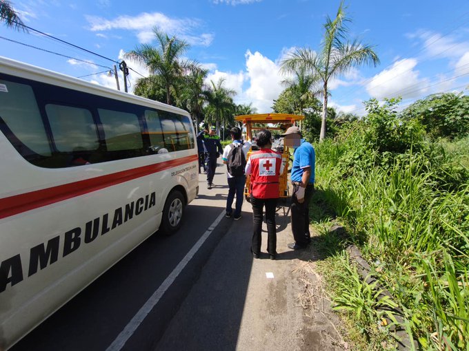 First image shows a white ambulance labeled Ambulancia parked on a roadside with palm trees and greenery, several people including those in red vests standing nearby discussing the scene. Second image depicts a female medic in red vest providing medical care inside the ambulance to a man with blurred face, using gloves and tools on a stretcher bed. Third image features a yellow tricycle involved in the accident, surrounded by police officers in uniforms, another ambulance, and bystanders near a water tank and fence under blue sky.