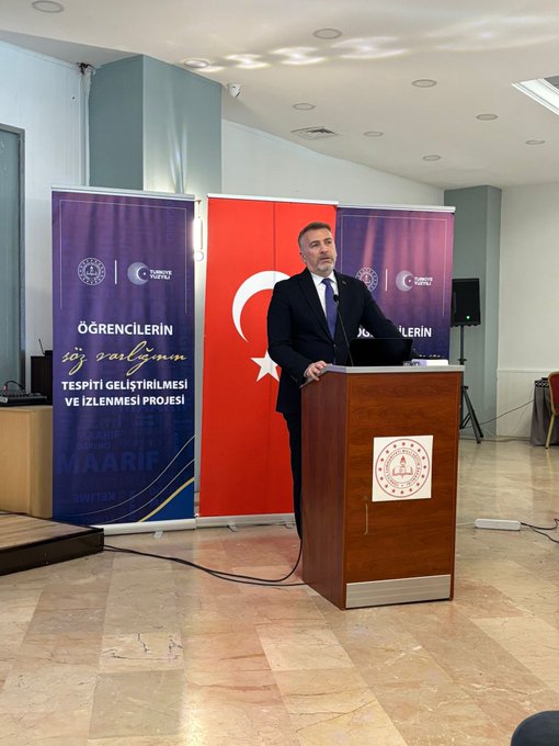 First image shows a man in a dark suit standing at a wooden podium speaking with a microphone in a modern conference room with blue and red banners displaying Turkish flags and text about student projects on either side light reflecting off the tiled floor and walls with office furniture nearby. Second image depicts a circular meeting setup with several professionals in business attire seated around a low table holding white flowers a projector screen displaying charts in the background a large portrait of a man on the wall and elegant lighting in a spacious room. Third image captures a diverse group of about 30 people including men and women in formal and semi-formal clothing posing together on wide stone steps outside a building with glass doors and autumn trees in the background under a clear sky.