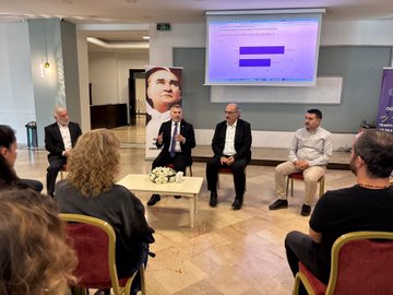 First image shows a man in a dark suit standing at a wooden podium speaking with a microphone in a modern conference room with blue and red banners displaying Turkish flags and text about student projects on either side light reflecting off the tiled floor and walls with office furniture nearby. Second image depicts a circular meeting setup with several professionals in business attire seated around a low table holding white flowers a projector screen displaying charts in the background a large portrait of a man on the wall and elegant lighting in a spacious room. Third image captures a diverse group of about 30 people including men and women in formal and semi-formal clothing posing together on wide stone steps outside a building with glass doors and autumn trees in the background under a clear sky.