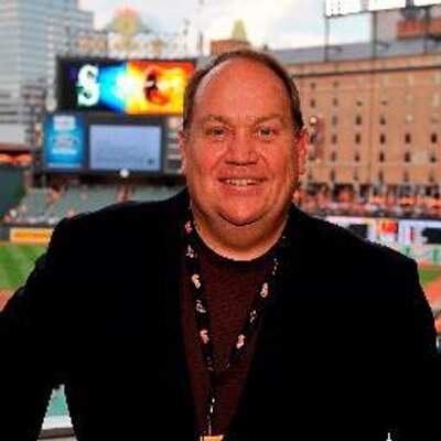 A man with balding hair and glasses stands smiling in a dark suit and red tie with a lanyard, positioned in front of a baseball field at night under stadium lights, with the Oriole Park scoreboard displaying colorful graphics in the background and city buildings visible.
