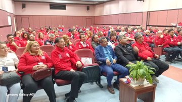 First image displays a large group of people mostly wearing red jackets with MEB AKUB logos standing together outdoors on steps near trees and a bench in an open area. Second image shows participants seated in rows inside a room with pink walls listening attentively some in red jackets others in suits around tables with plants and bags. Third image depicts a speaker standing at the front addressing an audience seated in a conference room with Turkish Ministry of National Education banners on the walls individuals in red jackets and casual attire focused on the presentation.