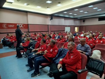 First image displays a large group of people mostly wearing red jackets with MEB AKUB logos standing together outdoors on steps near trees and a bench in an open area. Second image shows participants seated in rows inside a room with pink walls listening attentively some in red jackets others in suits around tables with plants and bags. Third image depicts a speaker standing at the front addressing an audience seated in a conference room with Turkish Ministry of National Education banners on the walls individuals in red jackets and casual attire focused on the presentation.