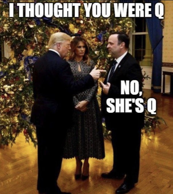Image depicts Donald Trump in dark suit gesturing with hand while speaking to a man in black suit and tie standing beside a decorated Christmas tree in an ornate room with blue curtains and wooden floor. Melania Trump stands to Trumps left in black patterned dress holding hands in front. Text overlay at top reads I thought you were Q in white font on black background, bottom text reads No shes Q with arrow pointing to Melania.