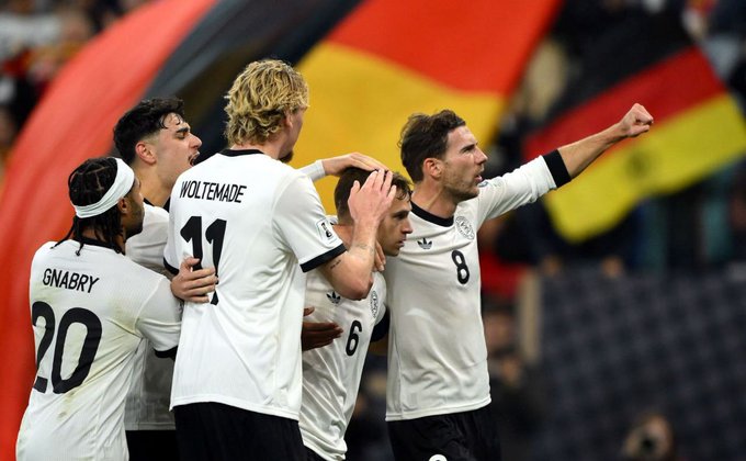 German national team players wearing white shirts with the numbers 20, 11, 6 and 8 celebrate together on the field in front of the black, red and yellow German flag. One of them raises his fist and another puts his hand on his teammate's head with the fans visible in the background.