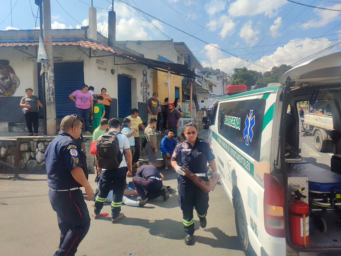 Street scene in a residential area with several emergency responders including police officers in blue uniforms and firefighters in yellow gear attending to a person lying on the ground near a white ambulance van marked with green stripes and a red cross, surrounded by onlookers and vehicles under a partly cloudy sky.